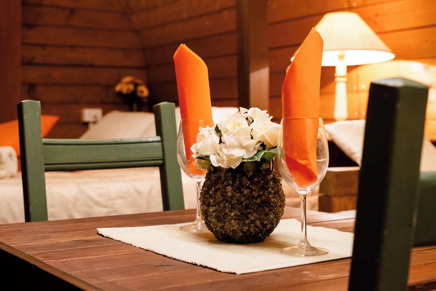 A wooden table with a glass vase containing white flowers and a pineapple, two glasses with orange napkins, and a lamp in a wooden room.