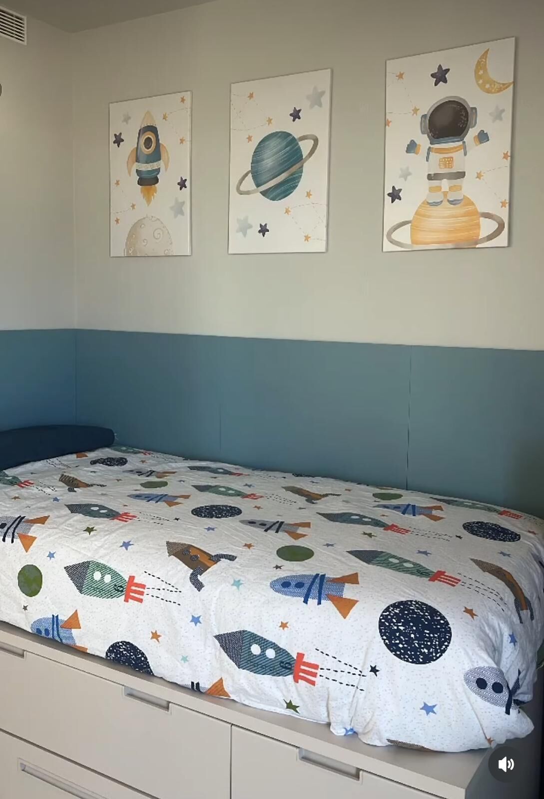 A bedroom with a bed featuring a space-themed bedspread, a blue headboard, and three wall-mounted space-themed paintings above the bed.