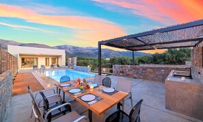 Un área de comedor junto a la piscina, con una mesa dispuesta para cuatro personas, rodeada de muros de piedra y una pérgola.