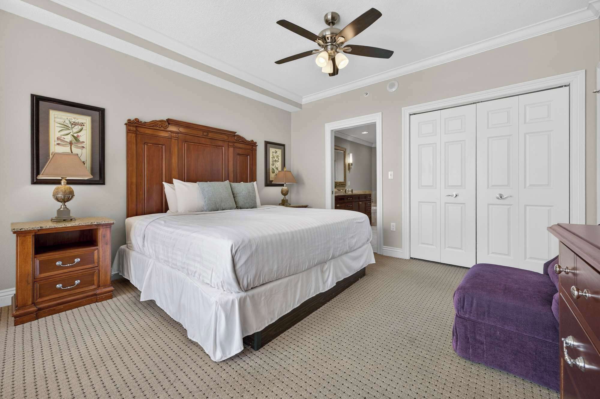 A bedroom with a large bed, wooden headboard, a ceiling fan, a dresser, a purple ottoman, and a white wardrobe.