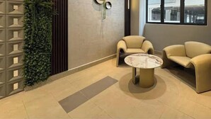 Um hall com uma mesa redonda e duas poltronas, uma parede decorativa com um relógio, e uma janela ampla.