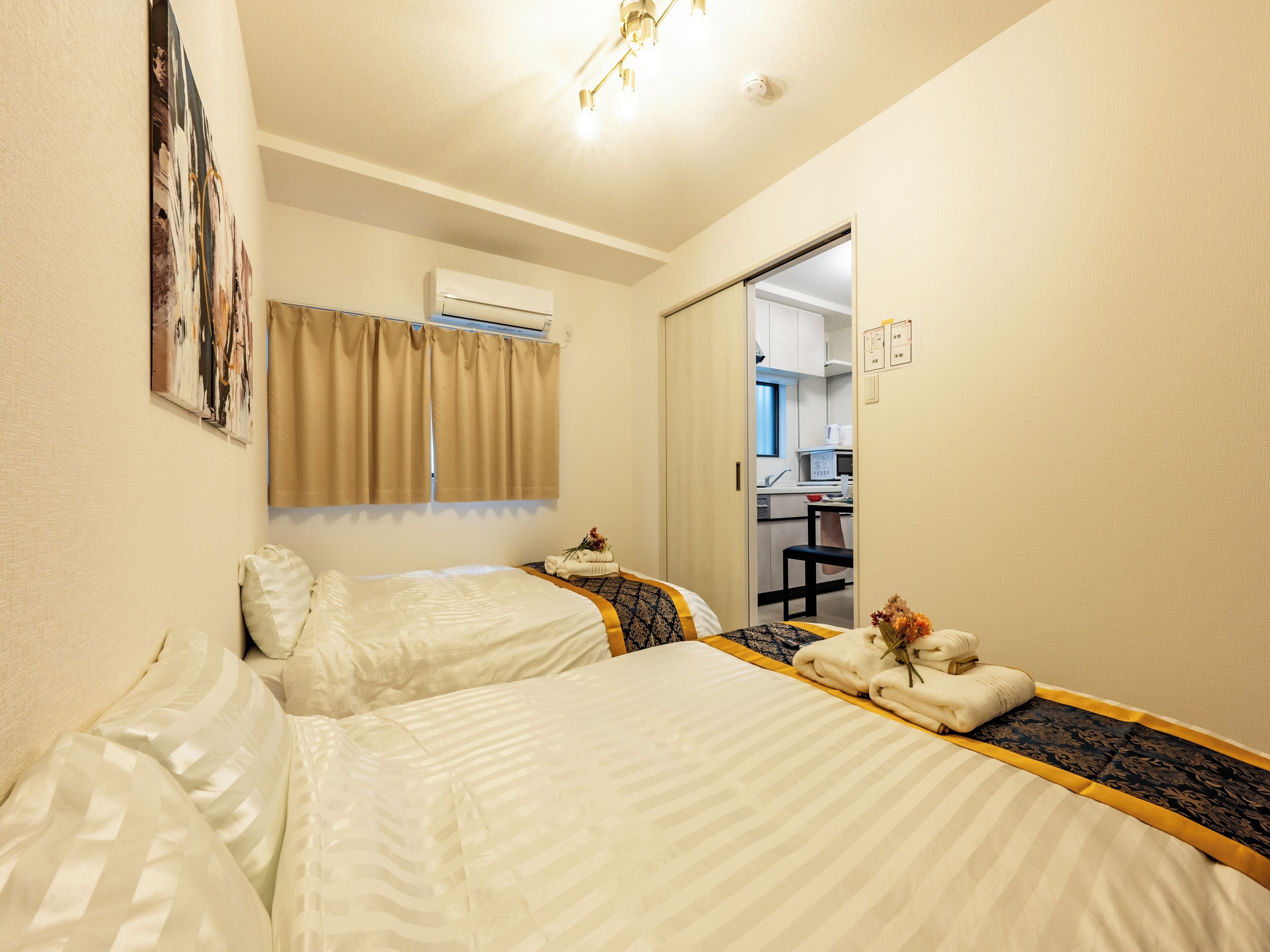 A hotel room with two beds, a window with curtains, an air conditioning unit, and a view into the kitchen area.