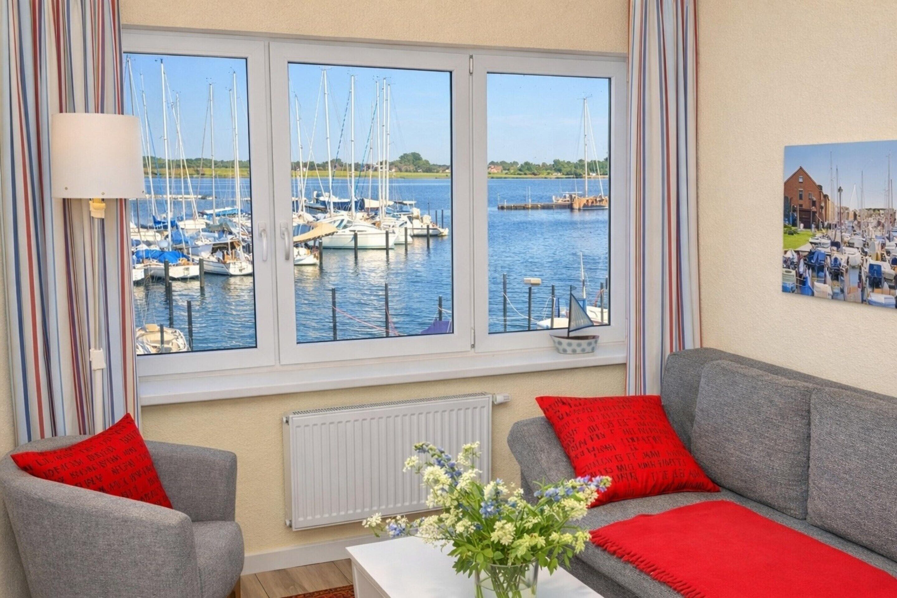 Una habitación con vista a un puerto deportivo, un sofá gris con cojines rojos, una mesa blanca y un jarrón de flores.