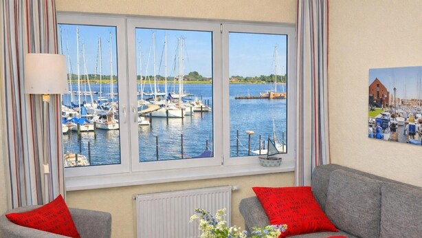 A living room with a view of a marina, a grey sofa, red cushions, a white table, and a vase with flowers. - Apartment Am Sudstrand (Fehmarn)