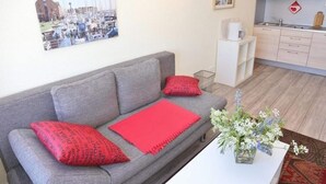 A living room with a grey sofa, red pillows, a red throw, and a vase with flowers. There is a kitchen area with a sink and cabinets. - Apartment Am Sudstrand (Fehmarn)