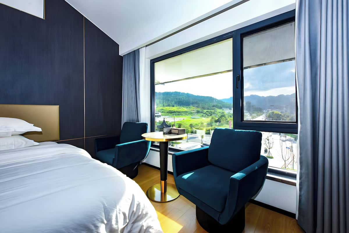 A modern hotel room with a large bed, two chairs, a small table, and a large window offering a view of a green landscape.