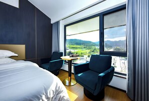Ett modernt hotellrum med en stor säng, två stolar, ett litet bord och ett stort fönster med utsikt över ett grönskande landskap.
