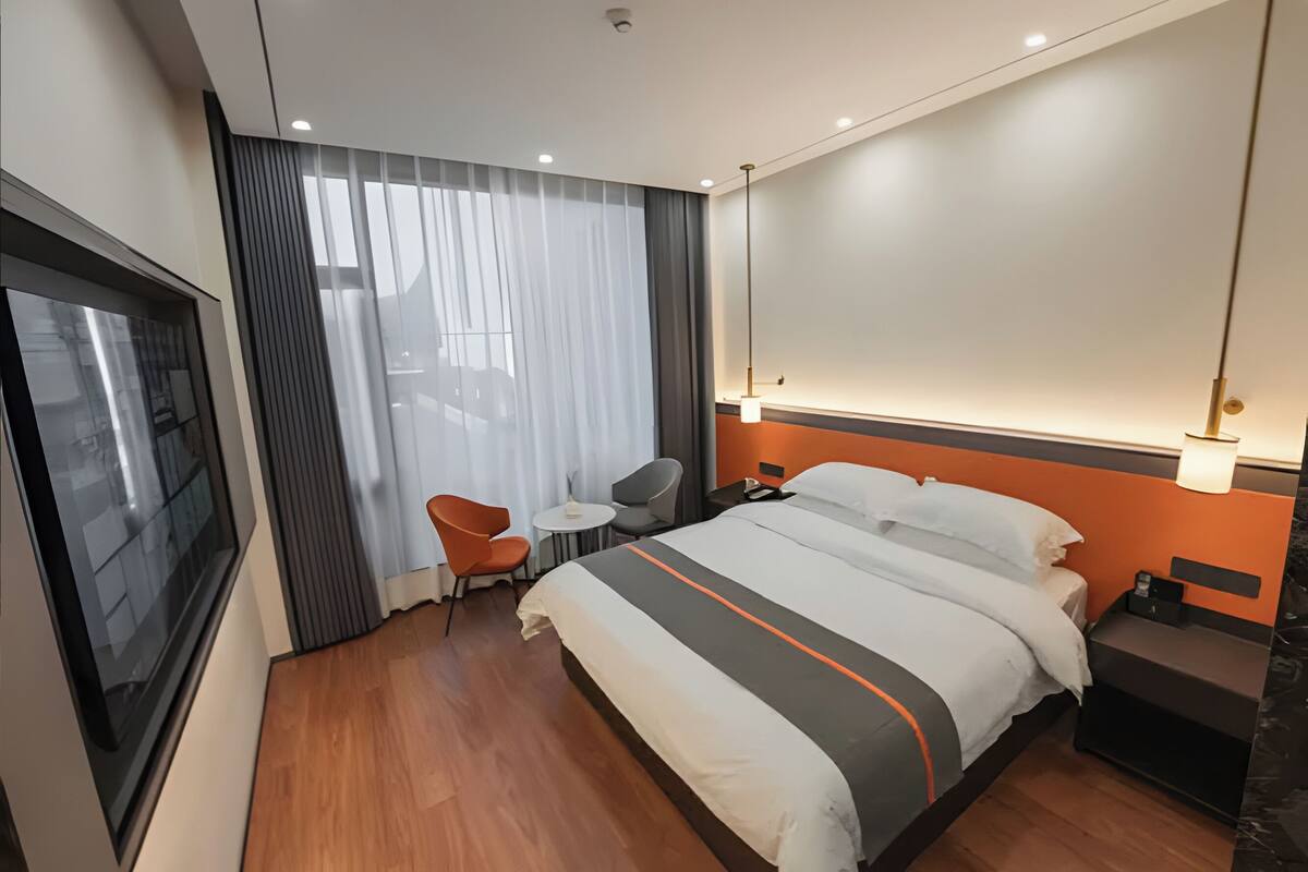 A modern hotel room with a large bed, a small table, a chair, and a large window with curtains.