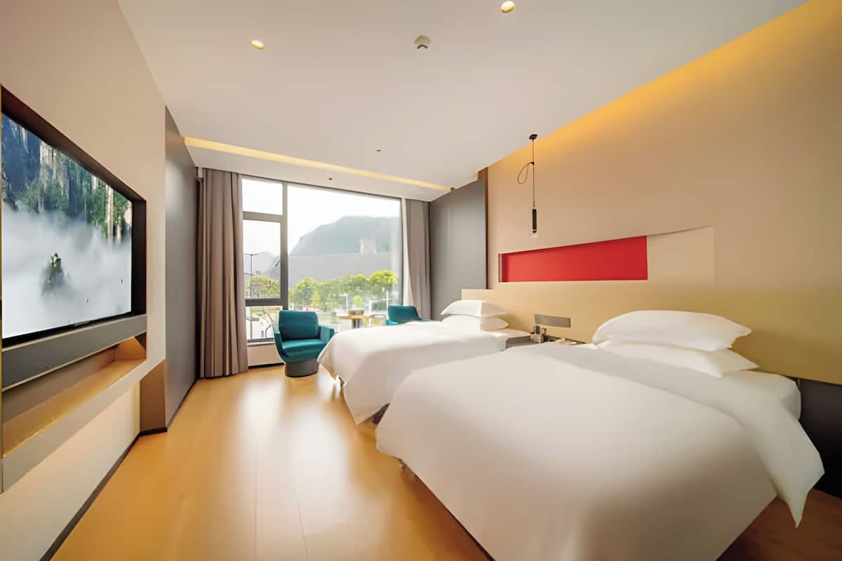 A hotel room with two beds, a TV, a large window with a view, and a red accent wall.