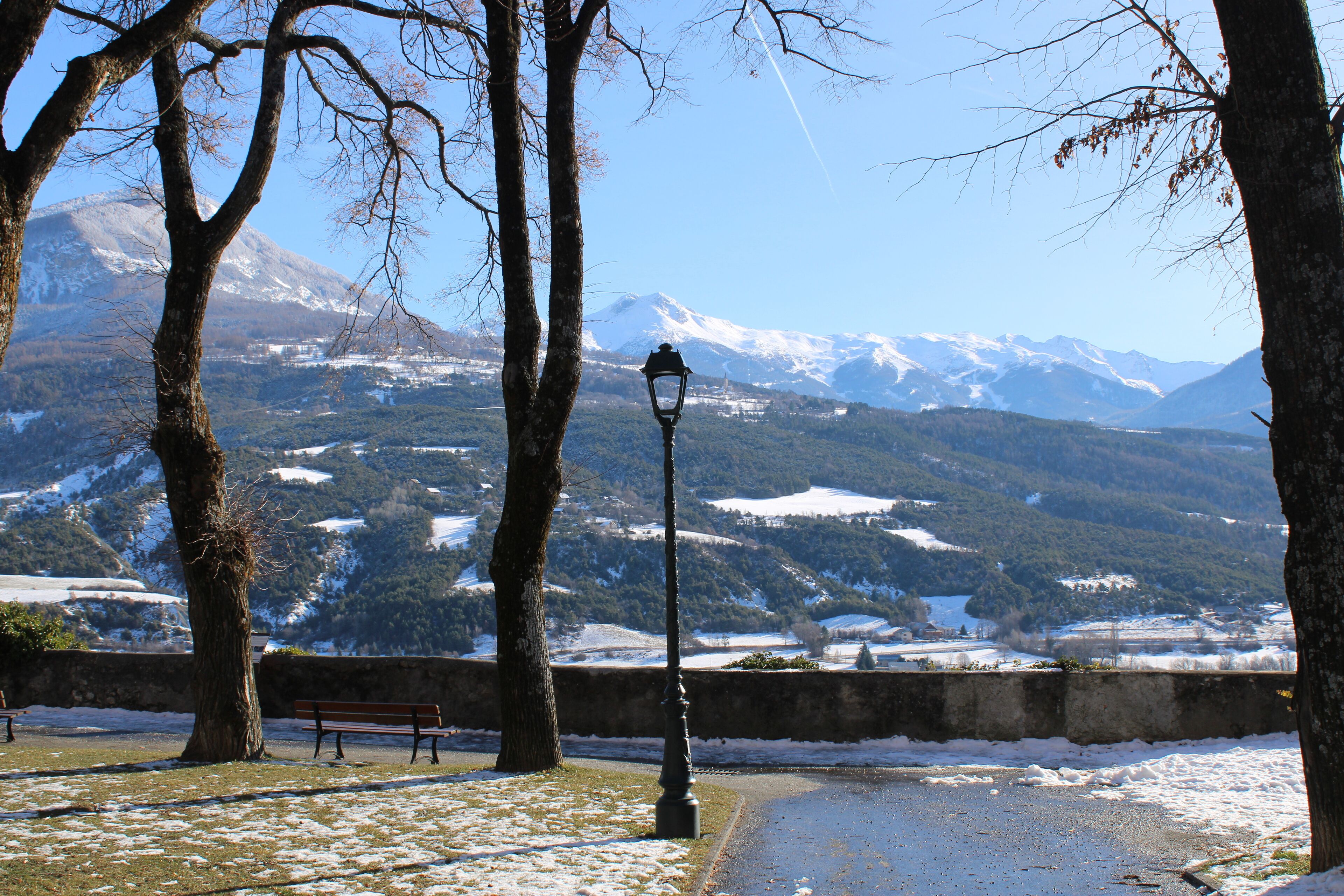 Eine Bank und ein Laternenpfahl in einem Park mit verschneiter Landschaft und Bergen im Hintergrund.