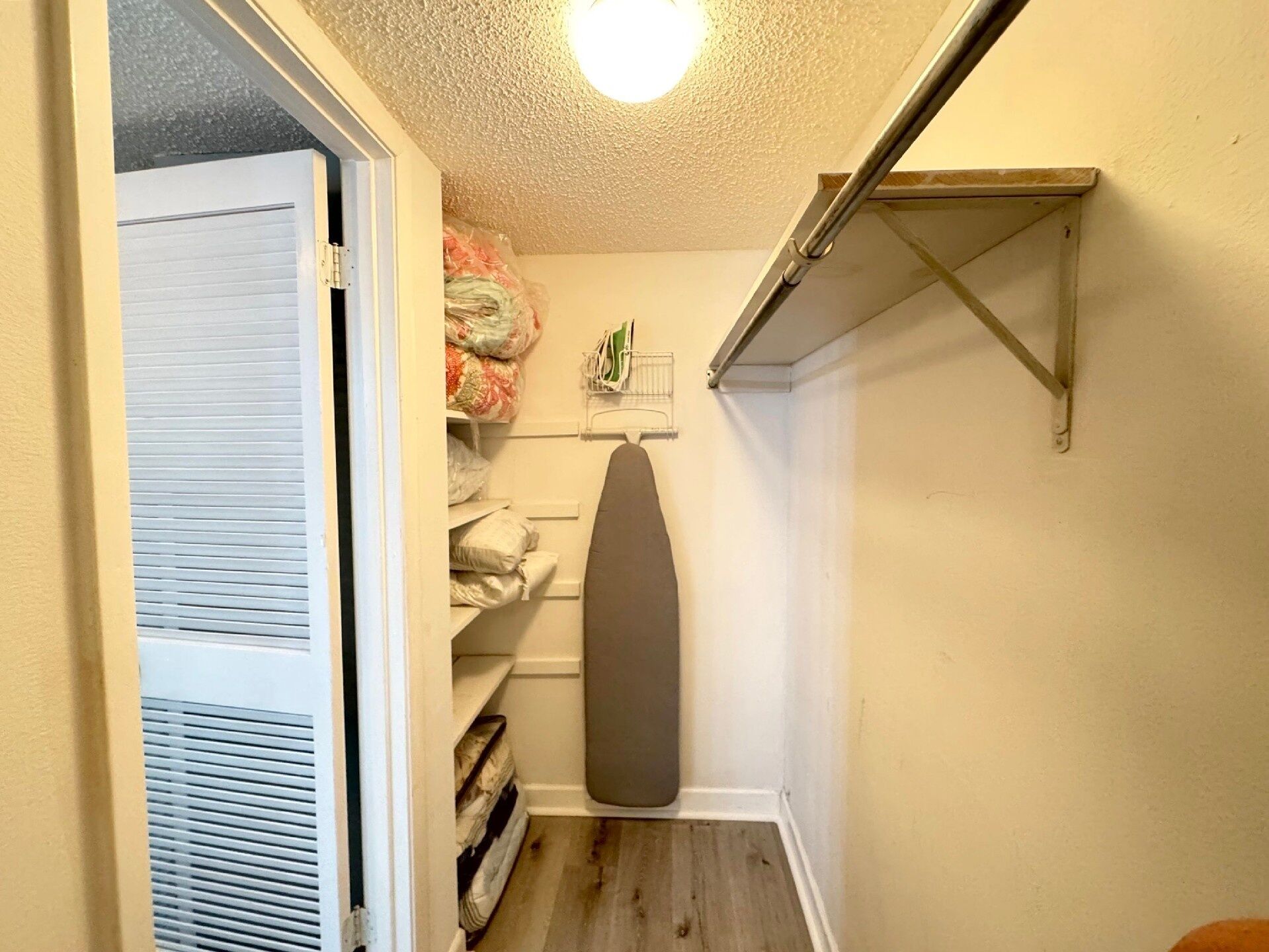 Pequeña lavandería con suelo de madera, una puerta blanca y un estante metálico con productos de limpieza.