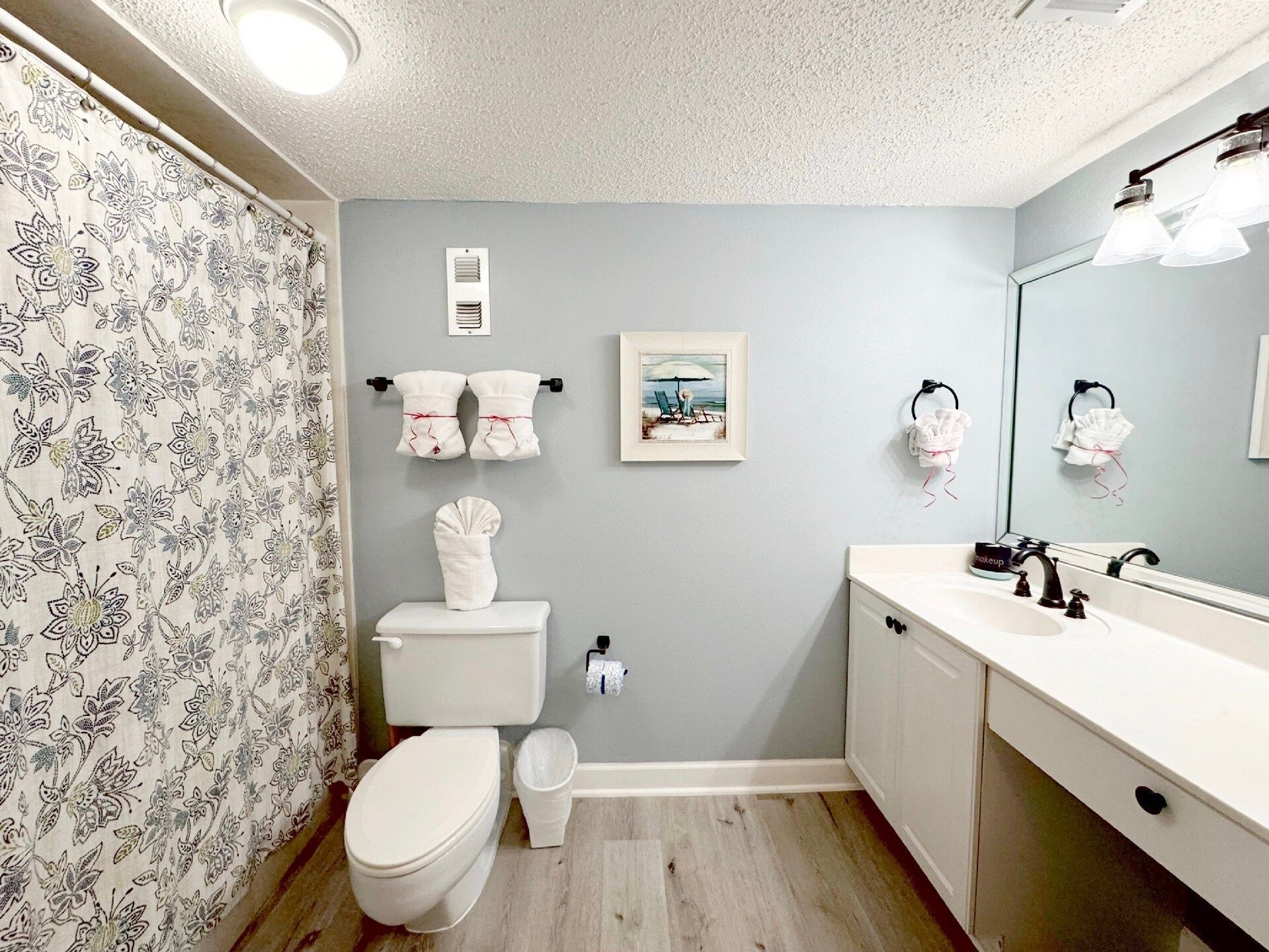 Une salle de bain avec des toilettes, un lavabo et un miroir. Des serviettes sont accrochées à des crochets et un tableau encadré est accroché au mur.