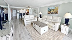 A spacious living room with a sofa, a coffee table, and a lamp. There is a kitchen in the background with a dining area. - Shoreline Towers 3103 2 Bedroom Condo (Destin)