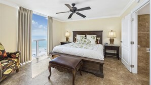 A hotel room with a large bed, two bedside lamps, a ceiling fan, a bench, a view of the beach, and a door leading to a bathroom. - Shoreline Towers 2075 2 Bedroom Condo (Destin)