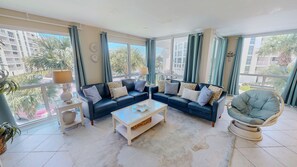 Ein modernes Wohnzimmer mit zwei dunklen Sofas, einem Glastisch und einem runden Sessel. Große Fenster bieten Blick auf Palmen und Gebäude.