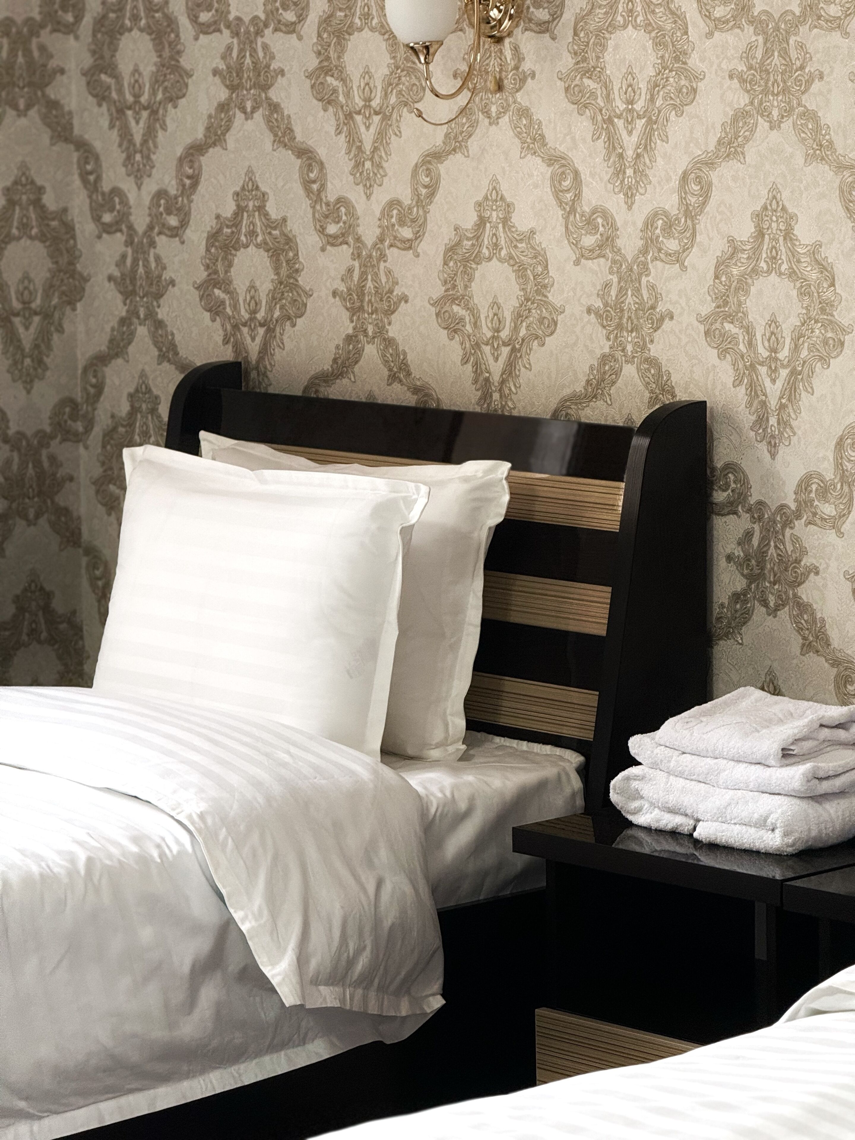 A neatly made bed with white linens and pillows, a wooden headboard, and a small bedside table with folded towels.