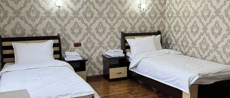 A hotel room with two single beds, wooden flooring, patterned wallpaper, bedside tables with lamps, and a ceiling light fixture.