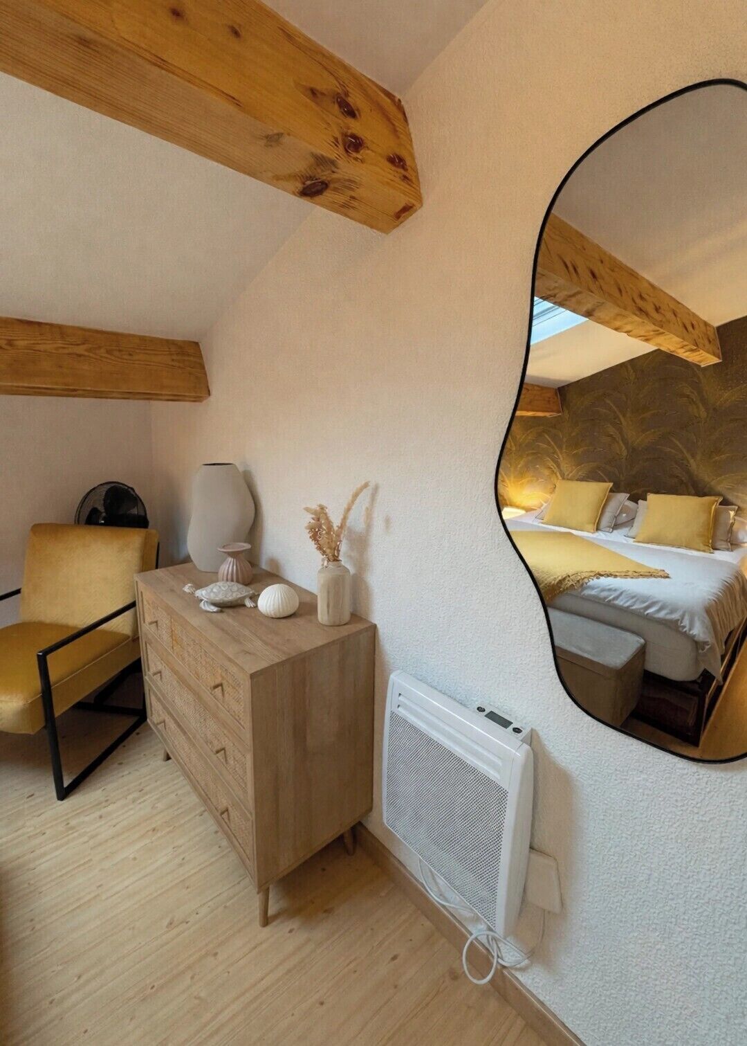 A bedroom with a wooden ceiling, a mirrored wall, a bed with yellow bedding, a bedside table with decorative items, a chair, and a radiator.