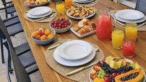 A dining table set with plates, bowls of fruit, and glasses of juice, surrounded by chairs and a potted plant.