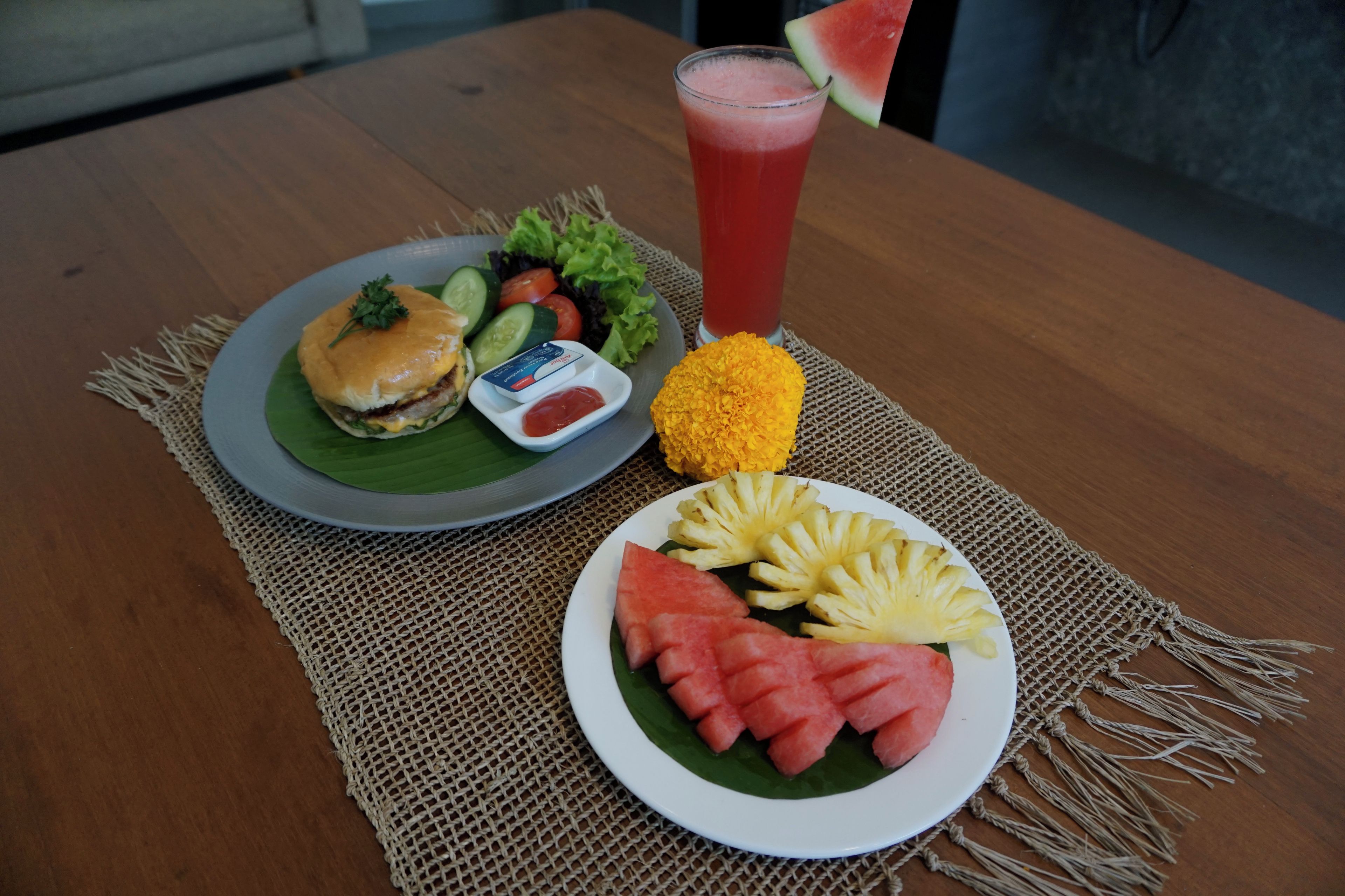 A meal consisting of a burger, salad, a glass of red drink, and fruit on a woven placemat.