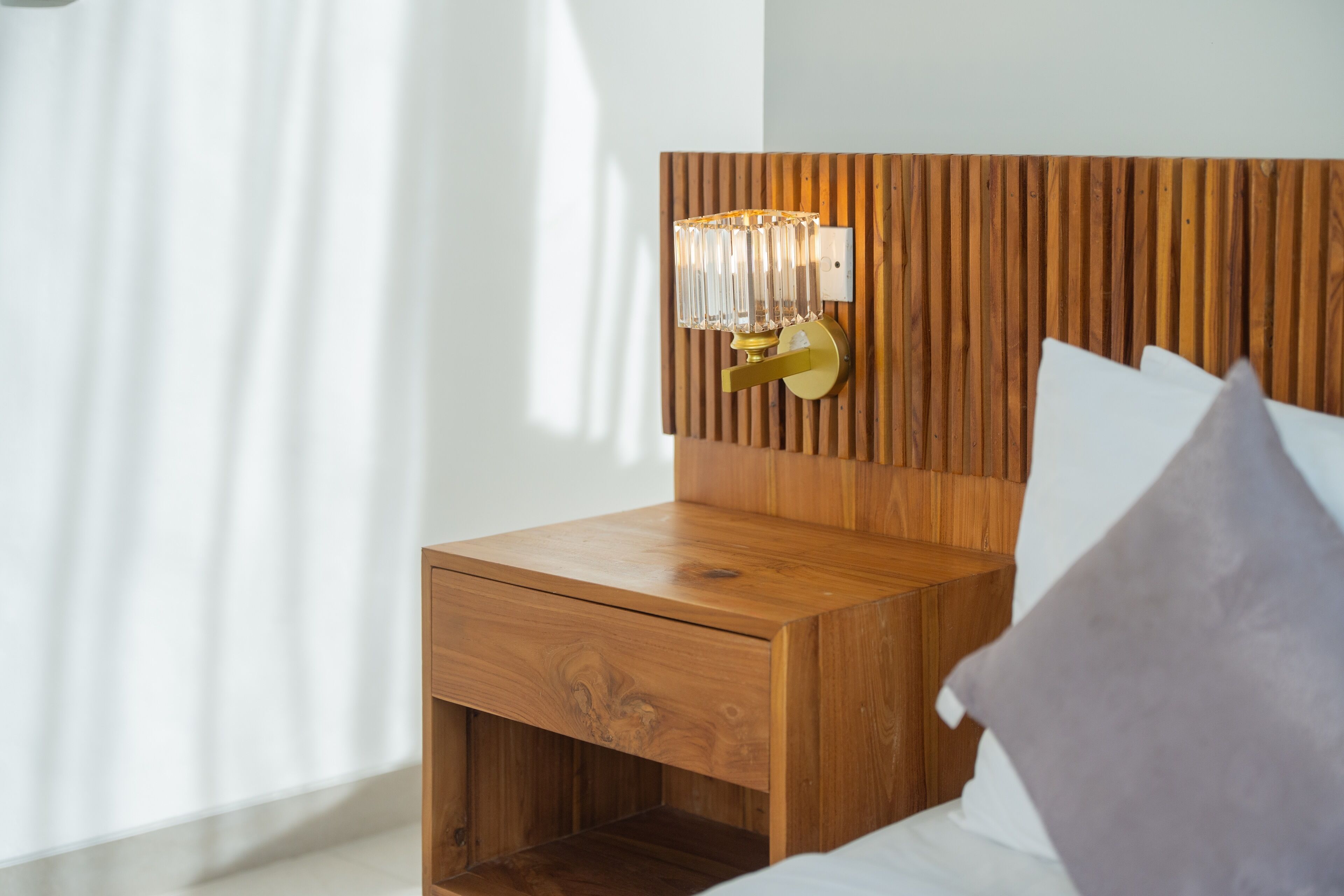 Uma mesa de cabeceira de madeira com uma gaveta, um candeeiro fixado na parede e uma cama com almofadas.