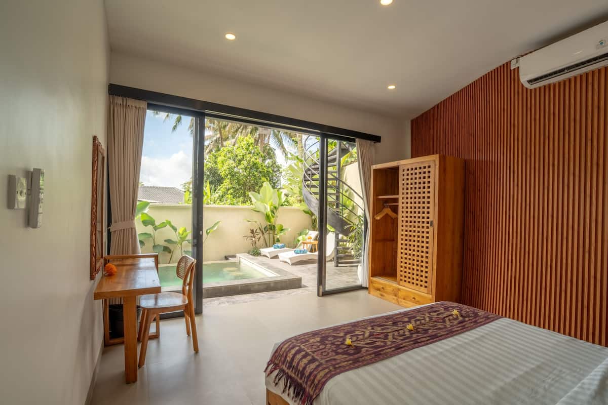 Un dormitorio con cama, escritorio, silla, armario de madera y puerta corrediza de vidrio que da a un patio con piscina y plantas.