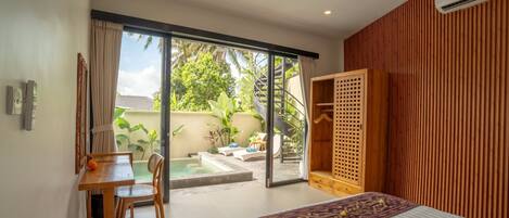 Ein Schlafzimmer mit Bett, Schreibtisch, Stuhl, Holzschrank und einer Glastür zum Patio mit Pool und Pflanzen.
