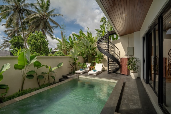Piscina com espreguiçadeiras, cercada por plantas tropicais e uma escadaria em espiral.