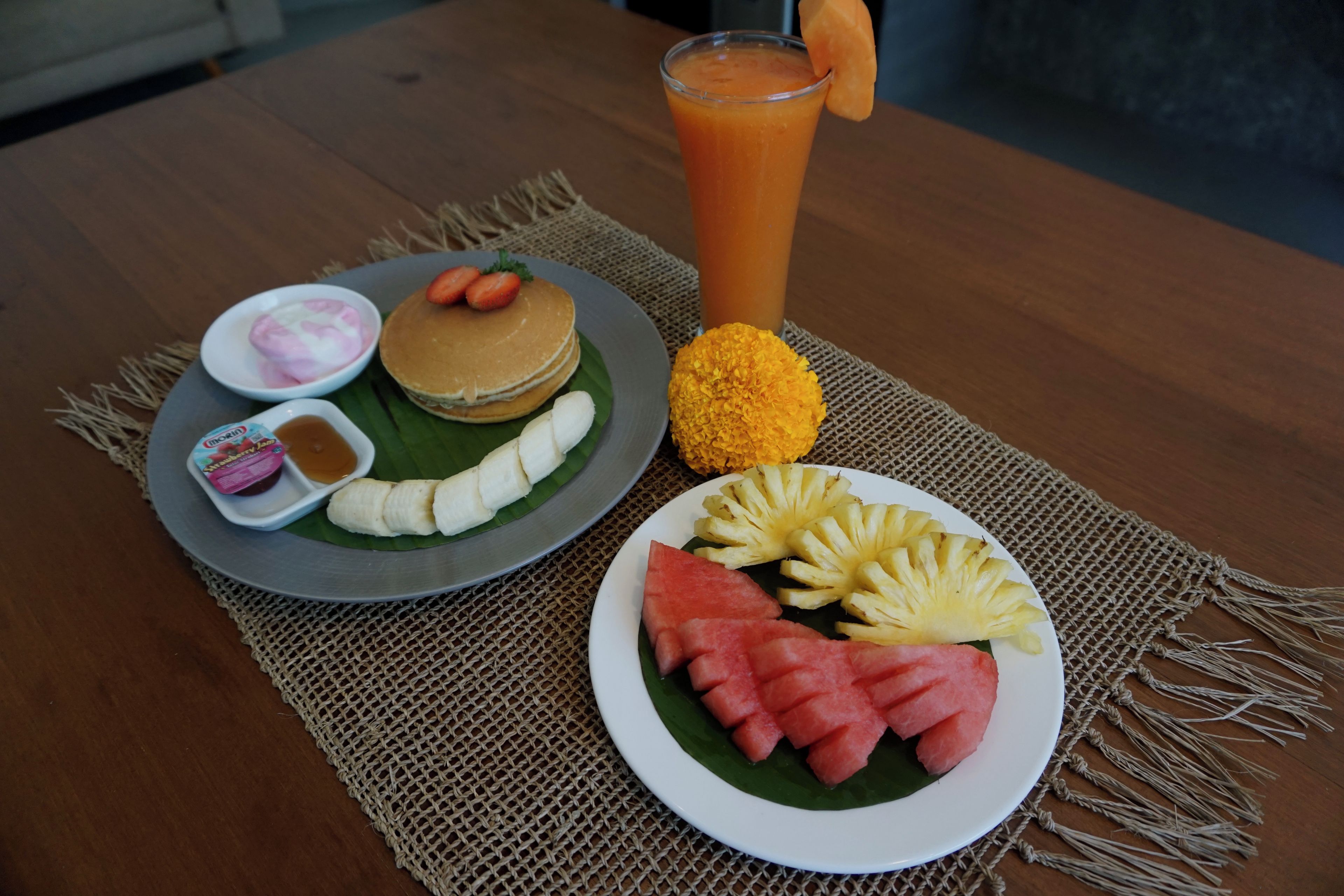 Une table dressée pour le petit-déjeuner avec des crêpes, des fruits, un verre de jus d’orange et un petit bol de yaourt.