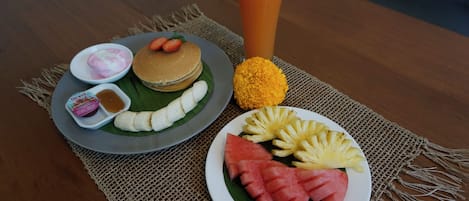 Une table dressée pour le petit-déjeuner avec des crêpes, des fruits, un verre de jus d’orange et un petit bol de yaourt.