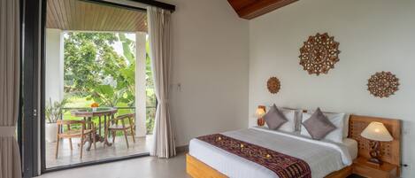 A bedroom with a large bed, a wooden nightstand, a table with chairs, and a view of greenery through a sliding door.