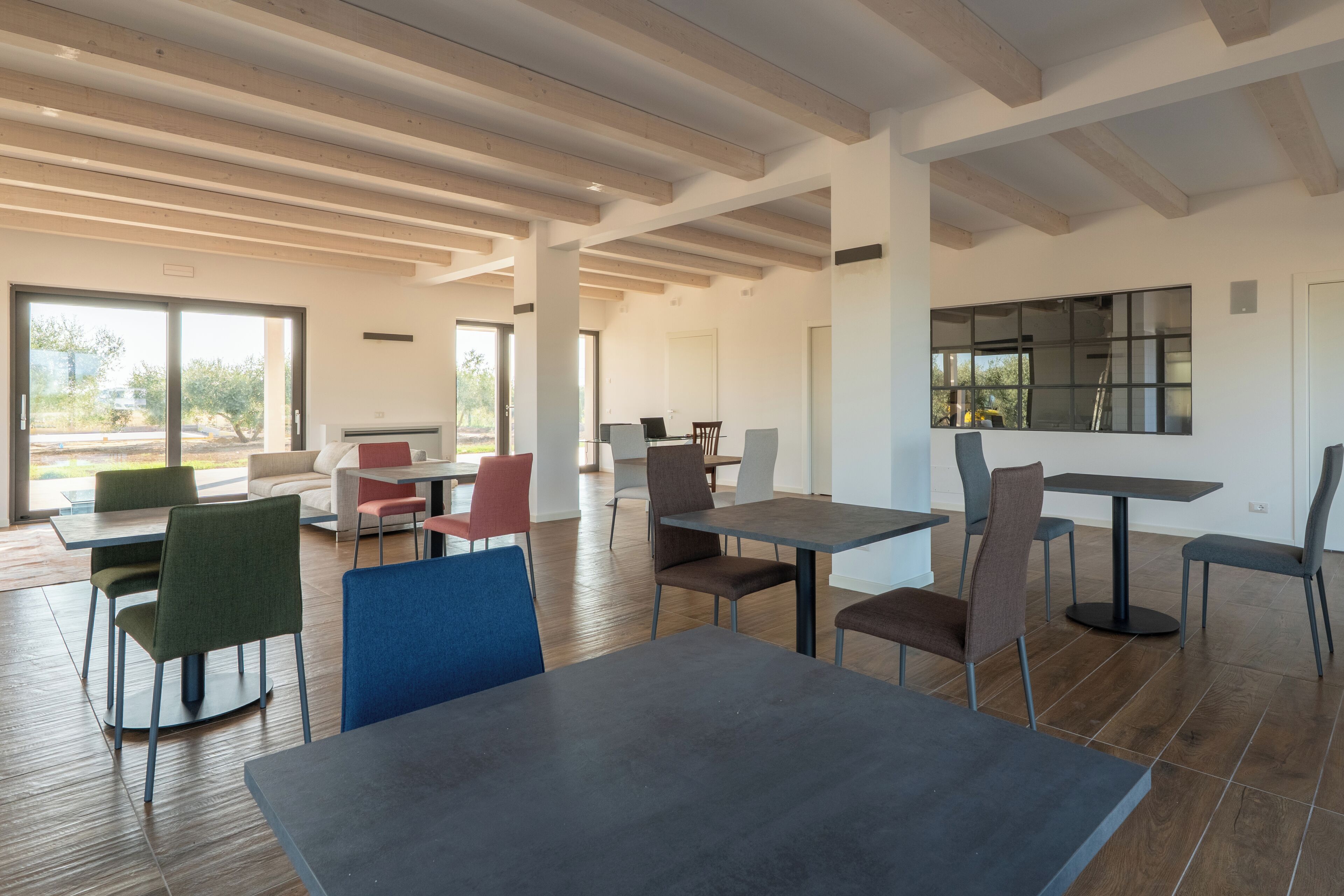 Un comedor moderno con vigas de madera, ventanas amplias y una variedad de colores en mesas y sillas.