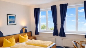 Ein Schlafzimmer mit Bett, Stuhl, Schreibtisch, Lampe, einem Fenster mit Vorhängen und einem Bild an der Wand.