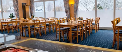 En buffet med forskellige spisemuligheder, herunder kød, oste og bagværk, i et rum med store vinduer og et flisebeklædt loft.