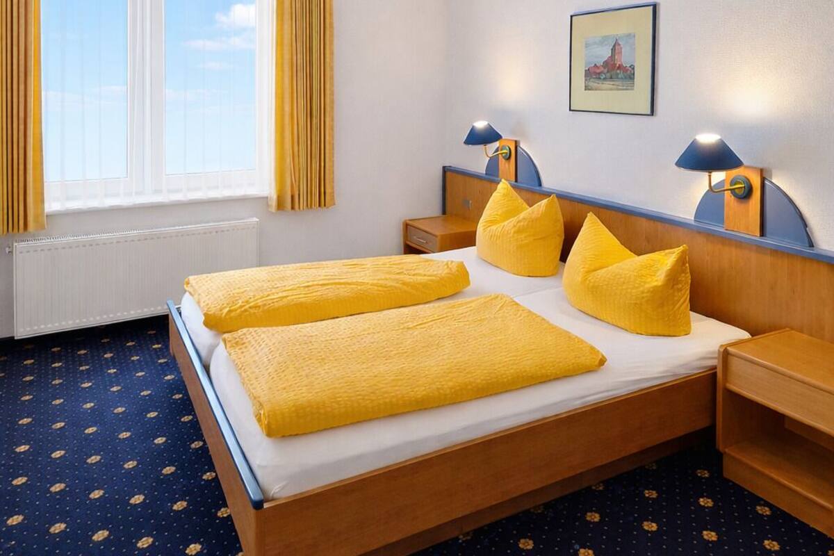 A hotel room with a double bed, two bedside tables, a window with curtains, a wall-mounted lamp, and a framed picture.