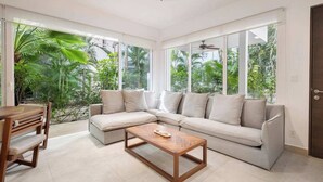 A modern living room with a sectional sofa, a wooden coffee table, and large windows offering a view of lush greenery. - Bright 2BR Condo | Aldea Zama (Tulum)