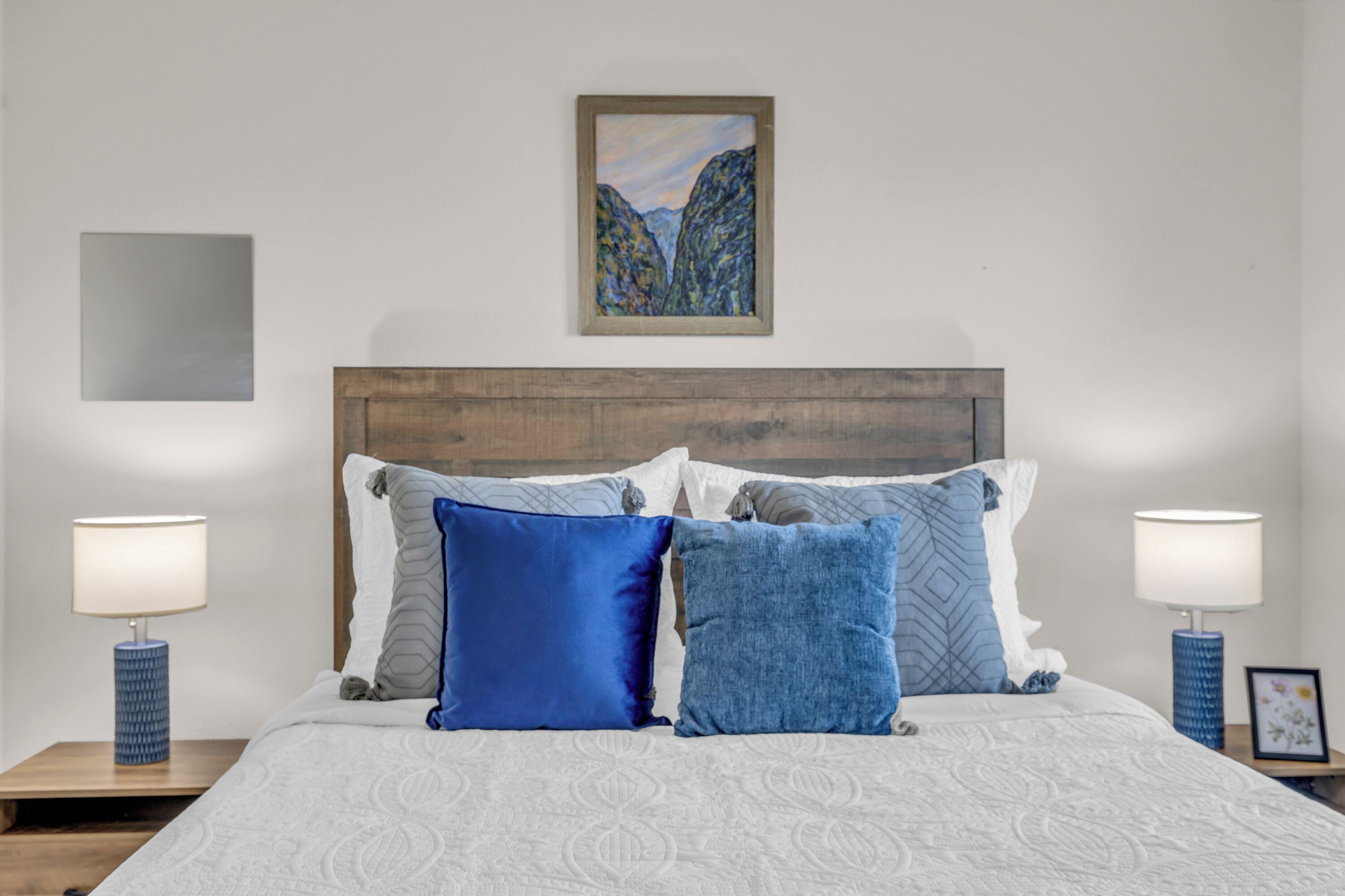 Una cama bien tendida con una colcha estampada, dos almohadas azules y un cabecero de madera. Encima de la cama hay un cuadro enmarco y, sobre la mesita de noche, una pequeña fotografía enmarcada.