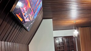 Een kamer met een aan een schuine muur bevestigde televisie, een houten plafond, een kroonluchter en een houten deur met ruitpanelen.