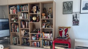 Uma sala de estar moderna com uma grande estante repleta de livros e objetos decorativos, um carro antigo vermelho e uma poltrona cinza com uma almofada colorida.