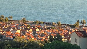 Uma vila costeira com um grande veleiro ao longe, rodeada por casas com telhados vermelhos.