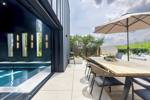 Area piscina esterna moderna con tavolo e sedie in legno, un ombrellone grande e vista sul verde circostante.