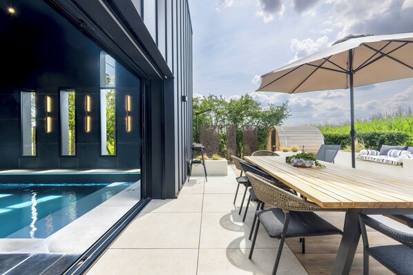 A modern outdoor pool area with a wooden table and chairs, a large umbrella, and a view of the surrounding greenery.