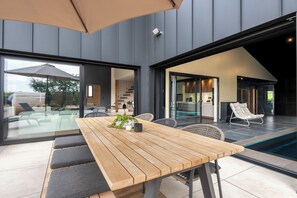 Un espace repas extérieur moderne, avec une table et des bancs en bois, entouré de portes vitrées donnant sur une piscine et un coin cuisine.