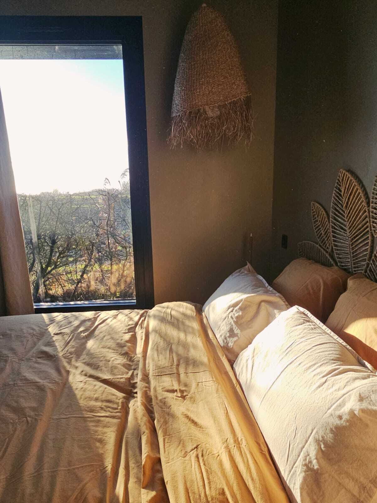 Une chambre avec une grande fenêtre, un lit recouvert d’une couverture à motifs et une tête de lit en osier.