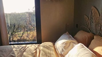 Une chambre avec une grande fenêtre, un lit recouvert d’une couverture à motifs et une tête de lit en osier.