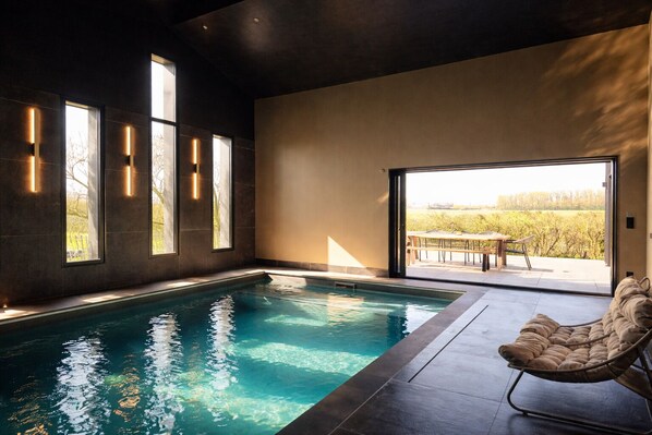 A modern indoor swimming pool with a view of a vineyard, a cushioned chair, and built-in lighting.