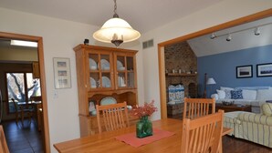 Un comedor con una mesa de madera, sillas y un florero. Se observa una pared de ladrillo y una pared azul con un sofá y almohadones.