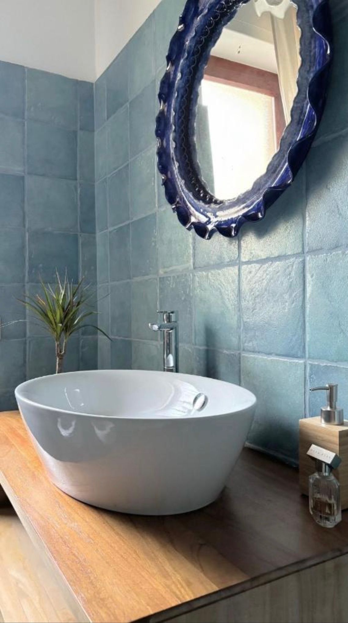 Une salle de bain avec un lavabo sur colonne blanc, un mur recouvert de carreaux bleus, un meuble-lavabo en bois et un miroir avec un cadre bleu.
