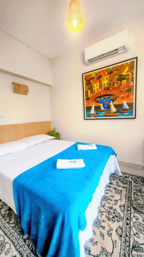 A hotel room with a bed, a blue bedspread, a painting of a coastal scene, and a patterned rug. - Eden Coworking Hotel - Pampulha  (Belo Horizonte)