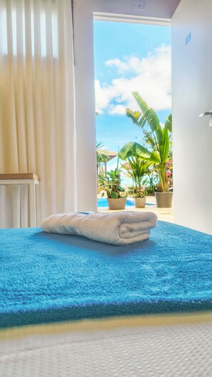 A room with a blue textured bedspread, a white towel on the bed, and a view of a pool and tropical plants through an open door. - Eden Coworking Hotel - Pampulha  (Belo Horizonte)